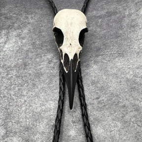 A close up photo of a  raven skull western bolo tie also known as a bola tie, string tie, or cowboy tie with a resin bird skull and braided cord made by Raven Ranch Studio.