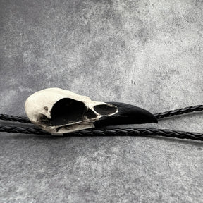 A side view photo of a raven skull western bolo tie also known as a bola tie, string tie, or cowboy tie with a resin bird skull and braided cord made by Raven Ranch Studio.