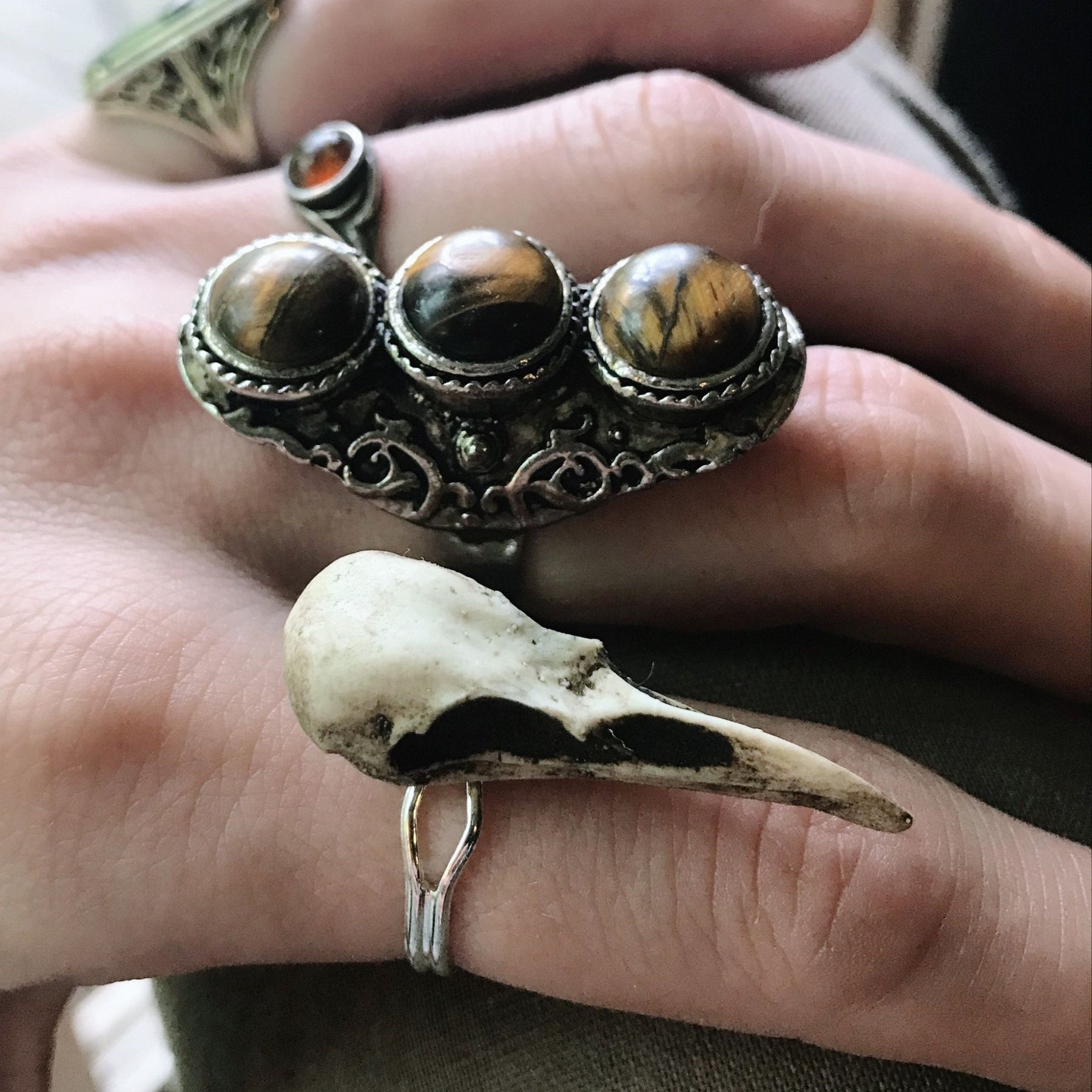Gothic bird skull witchy style resin raven skull ring on goth model hand next to silver and tiger's eye rings.