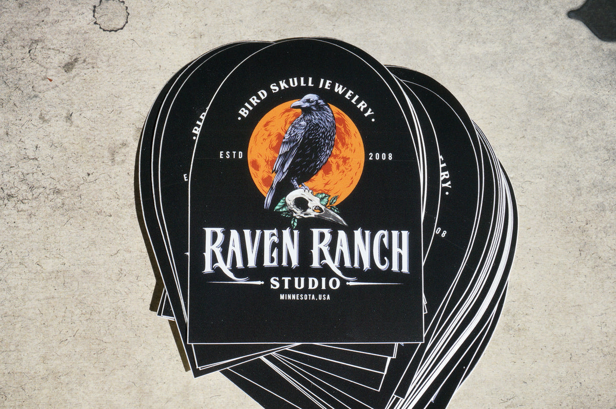 Stack of stickers for Raven Ranch Studio brand featuring a raven standing on a raven skull in front of a full moon.
